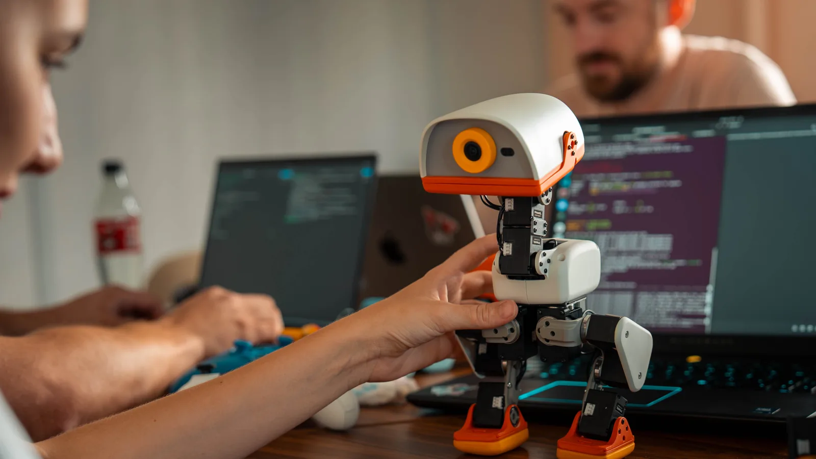 A hand steadying a cream-and-orange Microduck on a desk between two laptops running code, as taken from Pollen Robotics