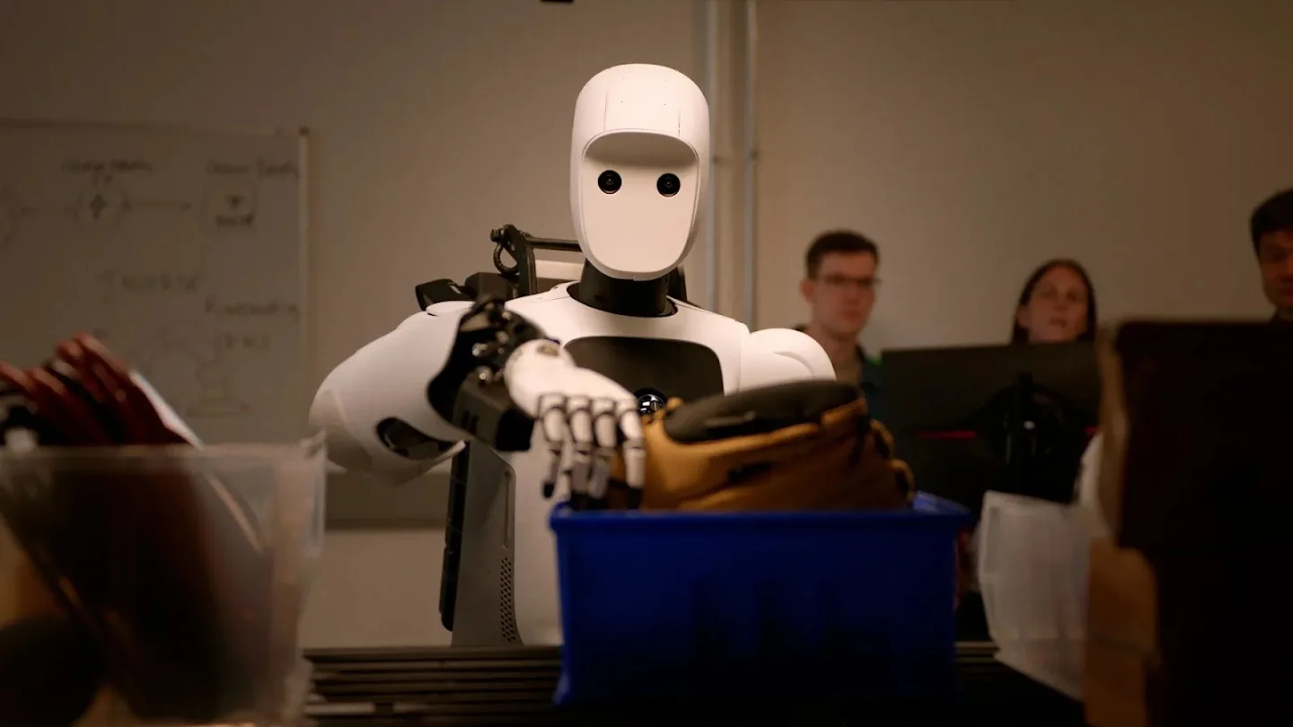 The Apptronik Apollo 2 humanoid reaching into a blue bin of fabric on a workbench while three researchers watch from behind laptops, as taken from Google DeepMind